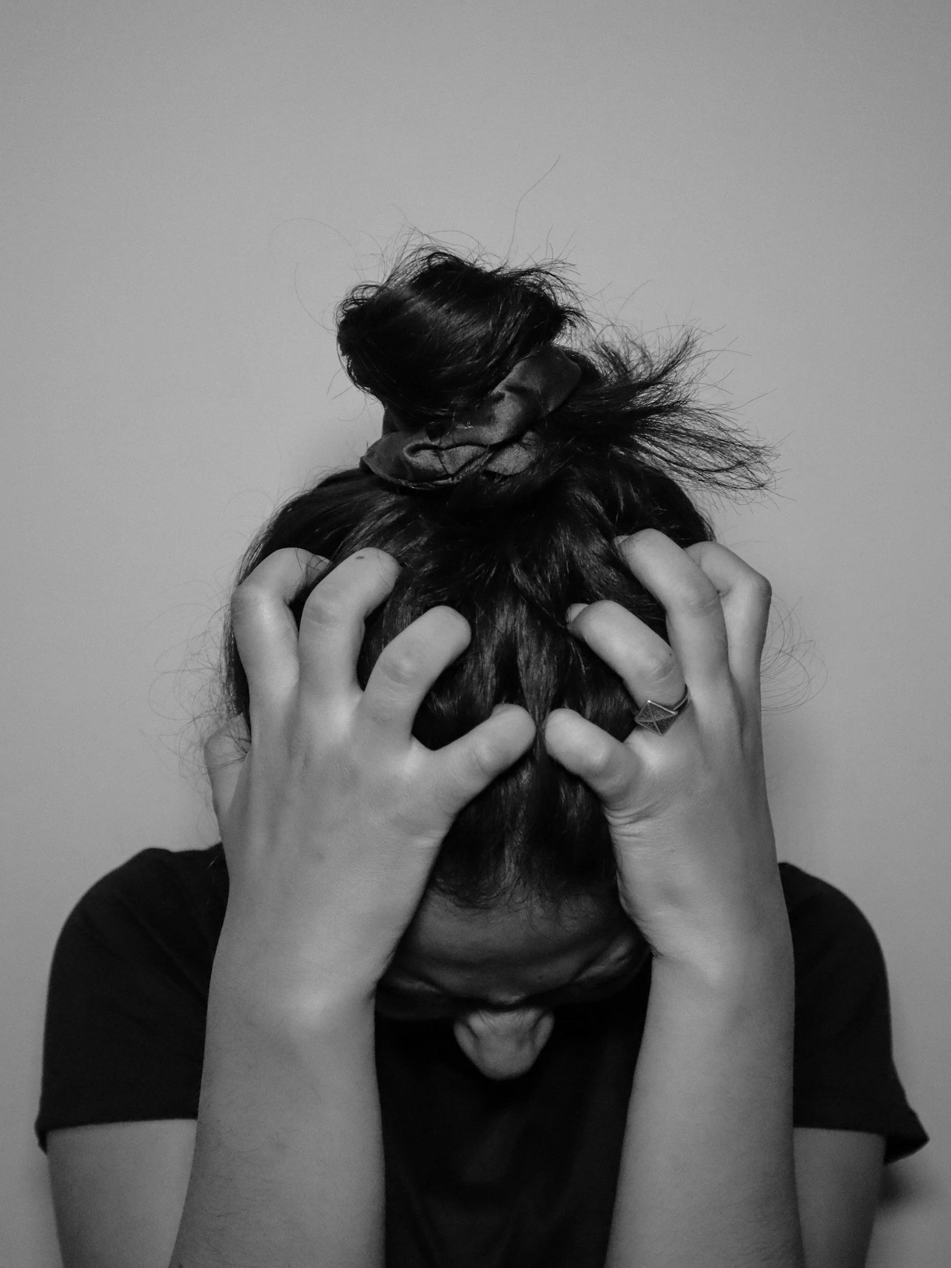 Person with long dark hair, wearing a black shirt, covering their face with their hands, with a loose bun on top of their head, black and white photo.